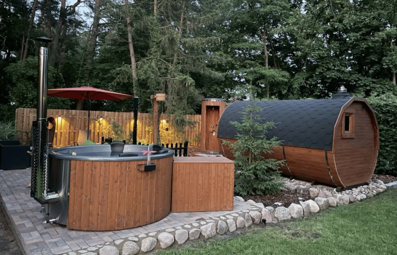Outdoor wellness area with wooden hot tub and barrel sauna in a landscaped garden, surrounded by trees and soft evening lighting