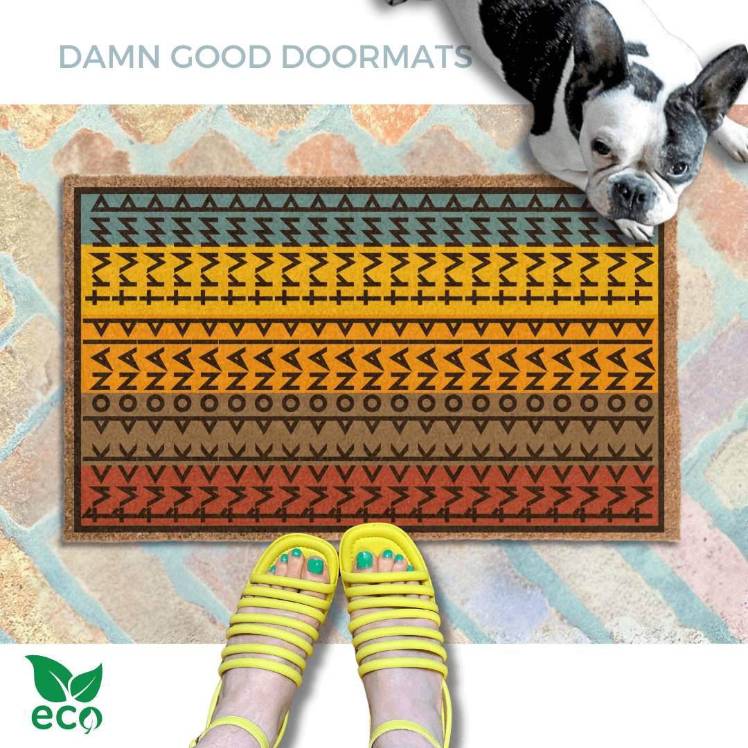a pair of feet in yellow strappy sandals standing on an all-weather colorful, patterned doormat with a geometric Fuck Donald Trump design and a french bulldog looking up.