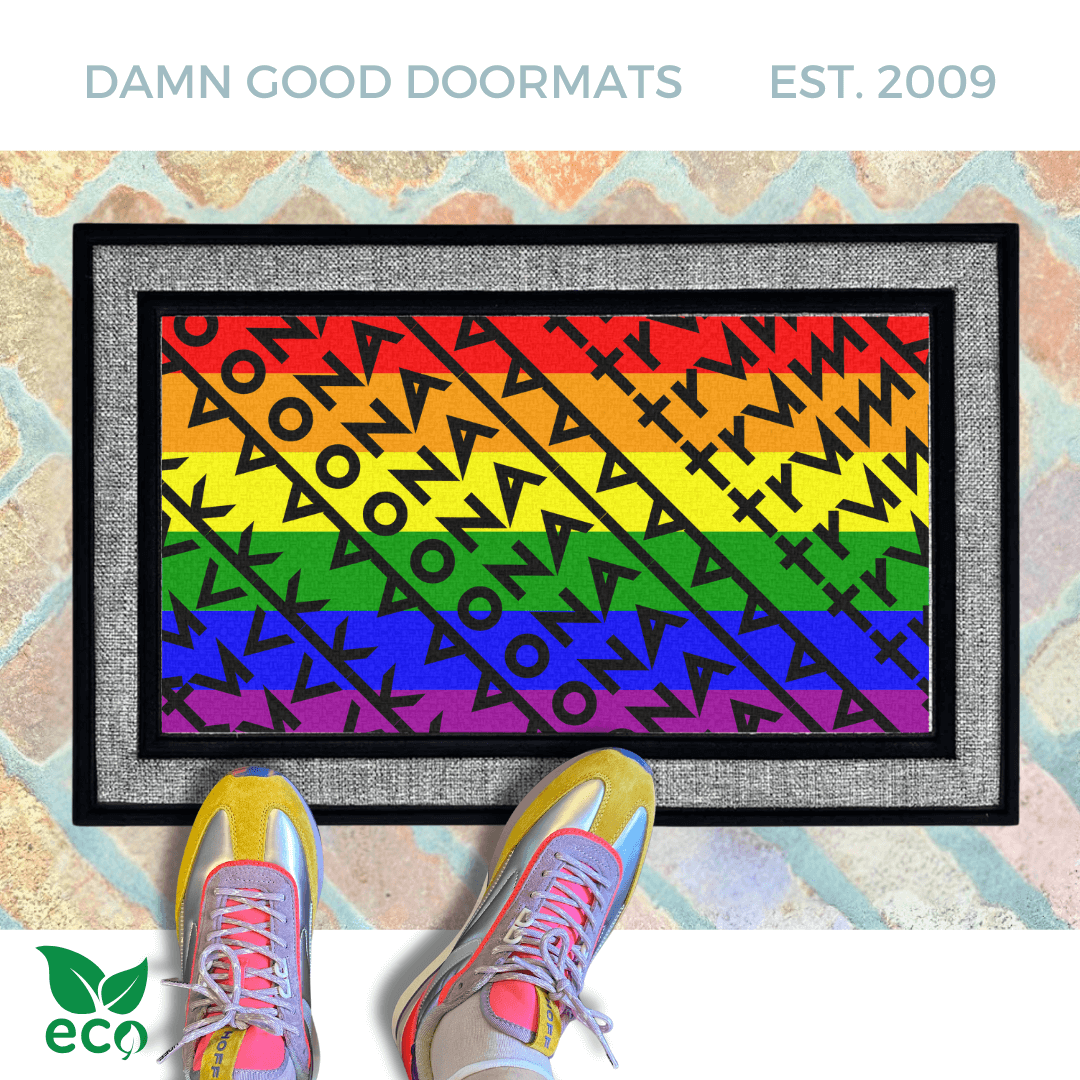 A gay pride rainbow-colored doormat patterned with a geometric design reading Fuck Donald Trump with a black pug dog standing on it, and a pair of bright neon colored sneakers next to it.