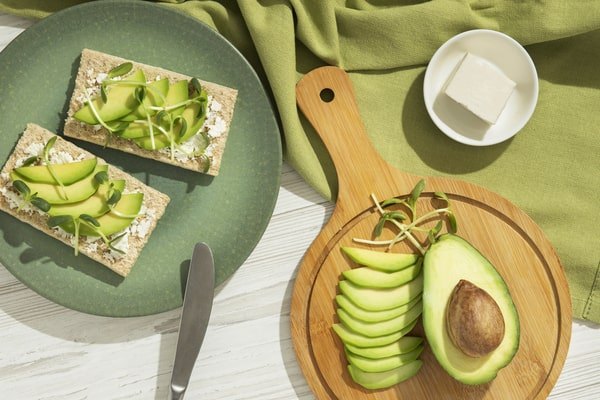 An overhead shot shows two avocado toasts topped with microgreens on a green plate next to a small wooden cutting board with a sliced avocado, a knife, and a square of feta cheese on a green cloth.
