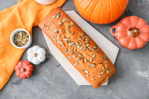 An overhead shot of a pumpkin loaf covered in pumpkin seeds on a white serving board, surrounded by decorative mini-pumpkins, a whole pumpkin, and an orange napkin.