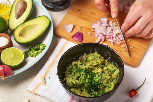 A pair of hands is chopping red onion on a small wooden cutting board, next to a black bowl of guacamole garnished with cilantro, and surrounding ingredients like halved avocados, limes, and garlic cloves.