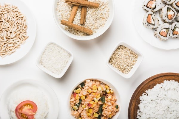 5 Keto Rice Substitutes With Fewer Than 5 Net Carbs 6 An overhead flat lay on a white background displays various rice products including puffed rice, uncooked long-grain rice, cooked white rice with a tomato slice, fried rice with vegetables, sushi rolls, and a bowl of uncooked rice garnished with cinnamon sticks.