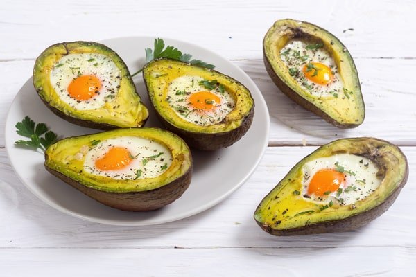 An overhead view shows five halves of baked avocado filled with a single egg, sprinkled with pepper and parsley, with three halves on a grey plate and two resting on a white wooden table.
