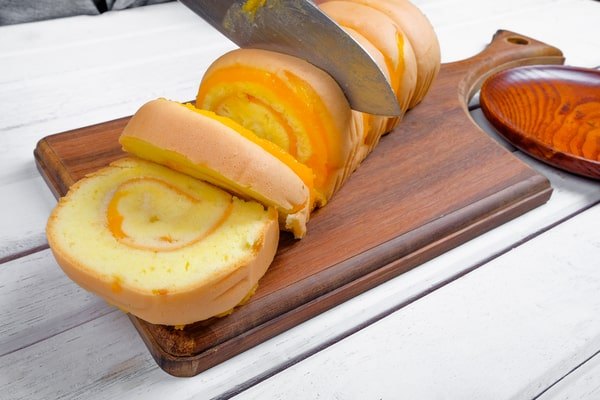 A knife is slicing a light yellow sponge roll cake with a pumpkin swirl filling on a dark wooden cutting board, resting on a white wooden plank table.